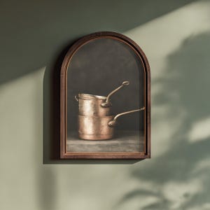 May include: A framed arched wall art piece featuring a dark, moody depiction of two stacked copper pots with long metal handles. The pots have a metallic, weathered finish and are positioned against a dark, textured background within a dark wood frame.