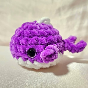 May include: A plush, purple crocheted whale with a white belly and a white loop. The whale has a black eye and purple fins. The soft, textured material gives the whale a cuddly appearance. This handmade item is a charming decorative piece.