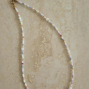 Könnte beinhalten: Eine zarte Perlenkette mit einer Vielzahl kleiner, mehrfarbiger Glasperlen in Weiß-, Rosa-, Gelb-, Blau- und Grüntönen, durchsetzt mit winzigen goldfarbenen Metallzwischenperlen und einem goldfarbenen Metall-Karabinerverschluss.