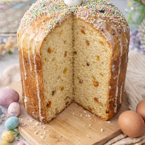 May include: A large, round Easter bread with a slice cut out, revealing a light-colored interior with dried fruit. The top is covered in colorful sprinkles and a white glaze, and a small bunny figure sits on top. The bread is on a wooden board.