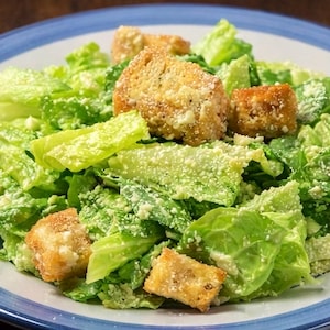 Può includere: Un'insalata Caesar servita su un piatto bianco con bordo blu, composta da foglie di lattuga romana verde tritata, crostini dorati tostati e un'abbondante spolverata di parmigiano bianco grattugiato su tutto il composto.