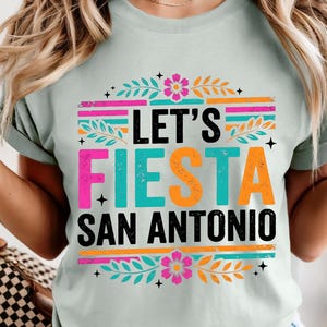 May include: A light green t-shirt with the text "LET'S FIESTA SAN ANTONIO" in a colorful design. The design includes pink, orange, teal, and black text with floral and striped accents. The shirt is short-sleeved.
