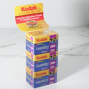 May include: Stacked boxes of Kodak Farbwelt 200 film with a yellow and red Kodak promotional display. The boxes are blue, yellow, and purple, with text in German. The display reads "100 Jahre Kodak in Österreich."