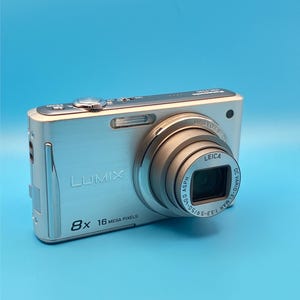Pode incluir: Uma câmara digital Panasonic Lumix prateada com zoom ótico de 8x, lente Leica DC Vario-Elmar 3.3-5.9/5.0-40.0 ASPH e resolução de 16 megapíxeis. O corpo da câmara tem um acabamento metálico com marca e texto claramente visíveis.