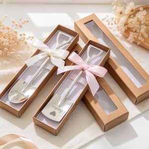 May include: Two silver-toned metal spoons shaped like miniature shovels, each engraved with a name and a heart icon. One spoon features a white ribbon bow, the other a pink ribbon bow. Both are presented in individual brown cardboard gift boxes.