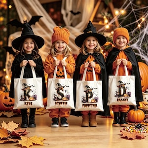 Puede incluir: Cuatro bolsas de tela blanca con letras temáticas de Halloween K, S, P y D, decoradas con fantasmas, calabazas y sombreros de bruja. Cada bolsa muestra un nombre: Kyle, Steffie, Preetvir y Dhanny, impreso en una fuente espeluznante y estilizada.
