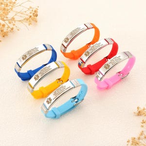 Könnte beinhalten: Sechs farbige medizinische Armbänder. Jedes Armband hat ein Silikonband in Blau, Orange, Rot, Pink, Gelb und Hellblau. Das Metallschild hat eingravierten Text und ein medizinisches Symbol. Der Text enthält Namen, Daten und medizinische Angaben.