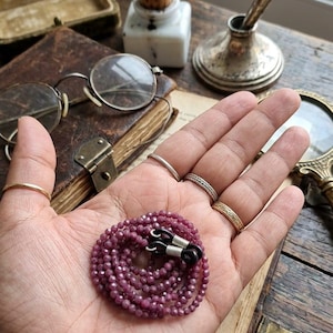 May include: A strand of small, faceted, deep pink beads forms a glasses chain. The chain has silver-toned connectors and black rubber loops for attaching to glasses. The chain is displayed on a hand, with other vintage items in the background.