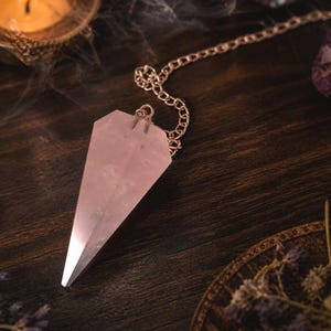 May include: A pink rose quartz pendulum with a silver chain hangs over a dark wooden surface. A lit candle and other crystals are in the background. The pendulum is a crystal used for divination and spiritual practices.