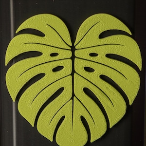 Könnte beinhalten: Ein schwarzer, sechseckiger Behälter mit einem großen, limettengrünen Monstera-Blatt-Design auf der Vorderseite. Das Blatt hat detaillierte Adern und eine strukturierte Oberfläche, wodurch ein 3D-Effekt entsteht. Das Design ist auf dem Behälter zentriert.