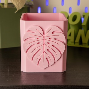 Puede incluir: Una maceta rectangular rosa claro con un gran diseño de hoja de monstera en relieve en la parte delantera. La maceta está sobre una superficie oscura. El fondo incluye una maceta verde y la palabra "plant" en verde.