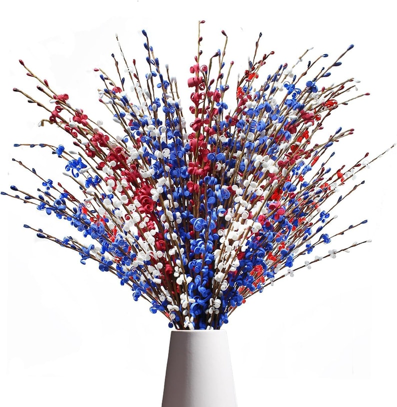 Op de afbeelding: Een boeket kunstbloemen in een witte vaas. De bloemen zijn rood, wit en blauw, wat een patriottisch kleurenschema cre&euml;ert. De bloemen zijn dicht op elkaar geplaatst en hebben een interessante textuur, ideaal voor decoratie.