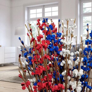 Op de afbeelding: Een decoratief bloemstuk met takken versierd met rode, witte en blauwe flessendoppen. De opstelling wordt binnenshuis gepresenteerd, met een neutrale achtergrond en natuurlijk licht. De flessendoppen zijn zo gerangschikt dat ze op bloemen lijken.