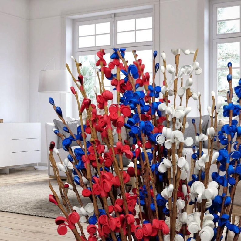 Op de afbeelding: Een decoratief bloemstuk met takken versierd met rode, witte en blauwe flessendoppen. De opstelling wordt binnenshuis gepresenteerd, met een neutrale achtergrond en natuurlijk licht. De flessendoppen zijn zo gerangschikt dat ze op bloemen lijken.