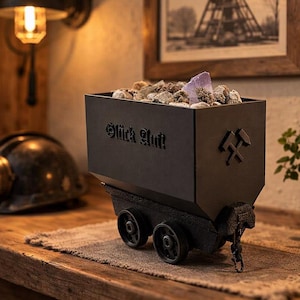 Pode incluir: Um carrinho de mina decorativo preto cheio de pedras e um grande cristal roxo. O carrinho tem as palavras "Glück Auf!" e um símbolo de martelo e picareta cruzados. Um capacete de mineiro vintage com uma lâmpada está por perto.