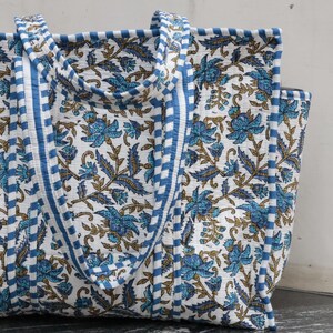Peut inclure: Un sac fourre-tout matelassé avec une base blanche ornée d'un motif répétitif de fleurs bleues et de feuilles vertes. Le sac comprend deux anses et des bordures décorées d'un motif à rayures horizontales bleues et blanches.