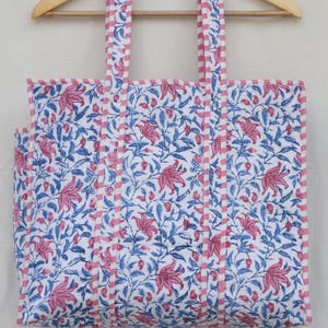 Peut inclure: Un sac fourre-tout rectangulaire à fond blanc orné d'un motif floral répétitif rose et bleu. Le sac présente deux anses verticales et des bordures en tissu rayé rose et blanc. La structure globale semble matelassée avec des coutures visibles.