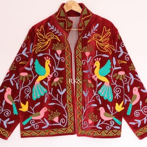 Op de afbeelding: Een donkerrood jasje met levendig, meerkleurig borduurwerk van vogels en bloemmotieven. Het ontwerp bevat gele, turquoise en roze vogels met groen en lichtblauw gebladerte. De binnenvoering heeft een wit en rood gestreept patroon.