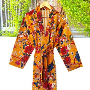 Peut inclure: Robe jaune moutarde ornée d'un motif floral rouge, bleu et blanc. La robe a des manches longues et une ceinture assortie. La doublure intérieure est blanche avec de fines rayures verticales. La robe est suspendue à un cintre en bois.