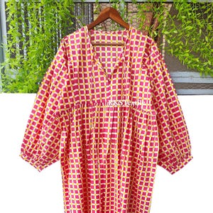 Puede incluir: Vestido de manga larga con estampado de cuadros rojos y amarillos. El vestido tiene un escote en V y una silueta suelta y fluida. La tela parece ligera, adecuada para climas cálidos. El vestido está colgado de una percha de madera.