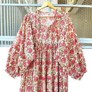 Puede incluir: Un vestido de manga larga con base blanca y un patrón repetitivo de flores rosas y hojas verdes. La prenda presenta un escote fruncido, una silueta holgada y puños elásticos en las mangas, todo ello expuesto en una percha de madera.