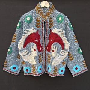 Könnte beinhalten: Hellblaue bestickte Jacke mit hohem Kragen. Die Jacke hat ein sich wiederholendes geometrisches Muster und ist mit roten Pferdeköpfen, floralen Designs und dem Text "RKS Textile" verziert.