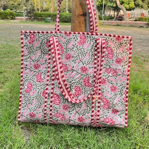 Può includere: Una grande borsa tote trapuntata con un motivo floreale rosa e verde su sfondo grigio. La borsa ha finiture e manici rossi. La borsa è appoggiata sull'erba verde.