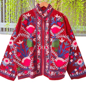 Puede incluir: Chaqueta roja bordada con cuello mandarín. La chaqueta presenta un diseño floral en hilo rosa, azul y blanco. El texto "RKS" es visible en la parte delantera. La chaqueta se muestra en una percha de madera.