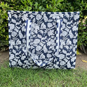 Op de afbeelding: Een rechthoekige gewatteerde tas met een donkerblauw en wit bloemenpatroon. De tas heeft twee bijpassende gewatteerde handvatten en een gestreepte rand langs de zijkanten. De stof toont een herhalende botanische print van bloemen en bladeren.