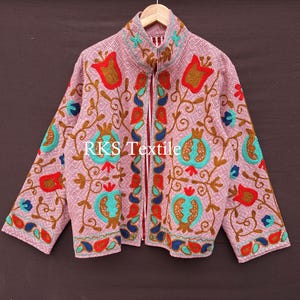 Op de afbeelding: Een roze jas met ingewikkelde bloemenborduurwerk in rood, turquoise en goud. De jas heeft een hoge kraag en een ritssluiting over de hele lengte. De tekst "RKS Textile" is zichtbaar op de voorkant. De jas hangt aan een houten hanger.