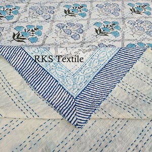 Puede incluir: Textil blanco y azul con estampado floral y borde a rayas azules. El textil presenta un diseño floral repetitivo en tonos azules y verdes. El texto "RKS Textile" es visible.