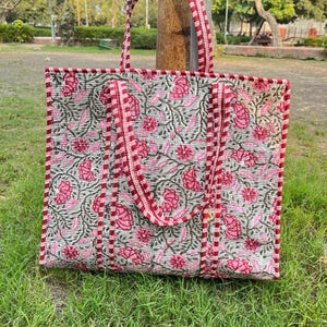 Puede incluir: Un bolso tote rectangular de tela acolchada con un estampado floral en tonos rosa y verde oscuro sobre un fondo gris claro. El bolso incluye dos juegos de asas y un ribete hecho de tela a cuadros rojos y blancos.