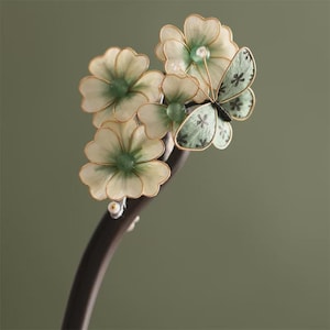 May include: A dark brown hair stick featuring a cluster of three cream-coloured flowers with green centres and a single light green butterfly with dark floral patterns on its wings. The floral and butterfly elements are accented with thin gold wire.