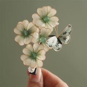 Könnte beinhalten: Eine Haarspange mit drei cremeweißen Blüten mit grünen Mitten und goldfarbenen Drahträndern, ergänzt durch ein kleines Schmetterlingsornament mit gemusterten grünen Flügeln. Die Blumen- und Schmetterlingselemente sind an einer dunklen Spange befestigt.