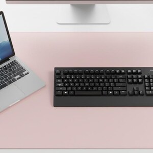 Pode incluir: Um tapete de mesa rosa claro com um laptop, teclado preto e mouse branco. O laptop exibe uma paisagem azul e branca. Uma pequena planta em vaso também é visível.