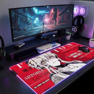 May include: A red and white rectangular gaming mousepad featuring a manga-style character illustration. The mousepad has a purple illuminated edge and the text "ARTORIA LANCER". A computer monitor, keyboard, and headphones are also visible.