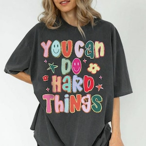 May include: Dark gray t-shirt with the text "You Can Do Hard Things" in colorful, bubble letters. The design includes smiley faces, stars, and flowers. The shirt has short sleeves and a relaxed fit.