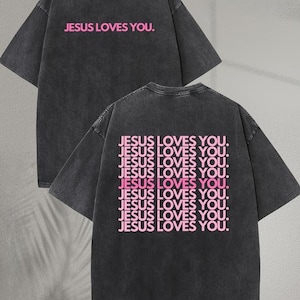 May include: Dark gray t-shirt with the text "JESUS LOVES YOU" in pink on the front and back. The back of the shirt repeats the phrase multiple times. The shirt has a crew neck and short sleeves.