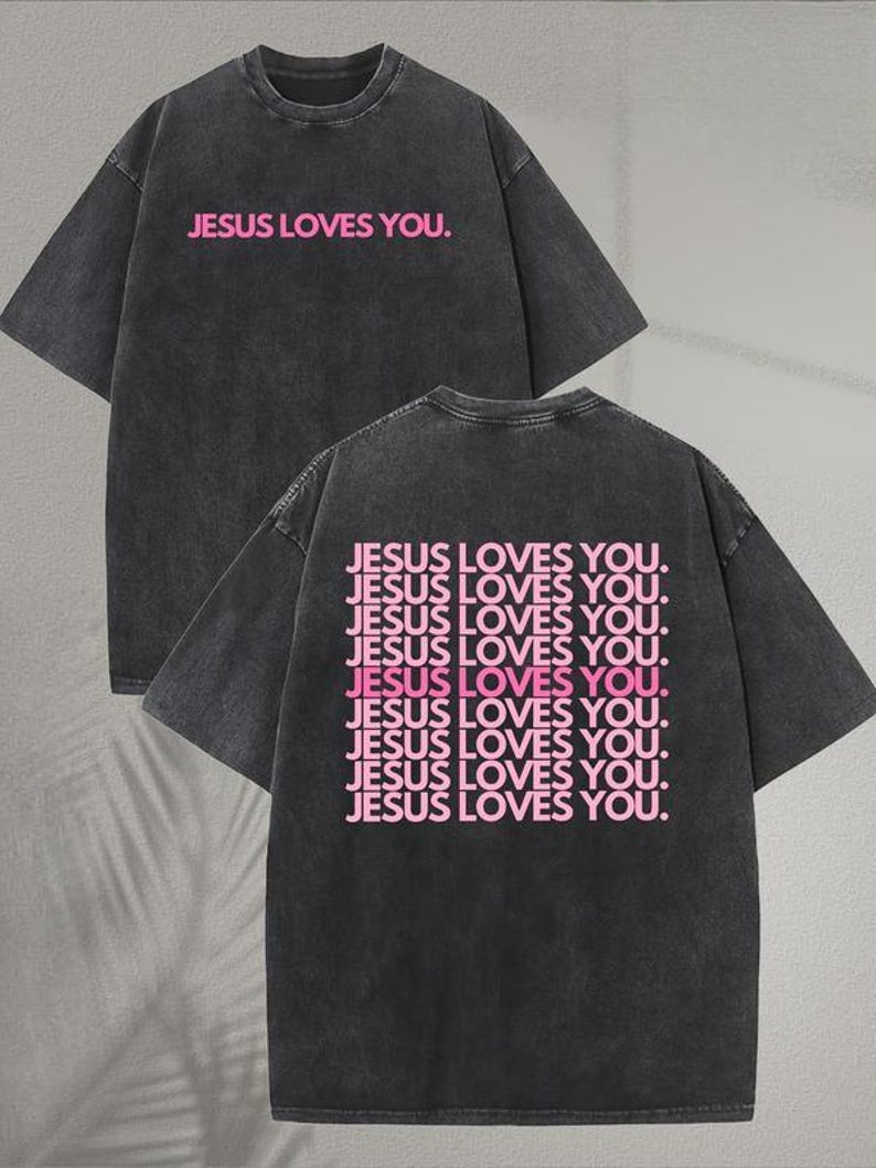 May include: Dark gray t-shirt with the text "JESUS LOVES YOU" in pink on the front and back. The back of the shirt repeats the phrase multiple times. The shirt has a crew neck and short sleeves.