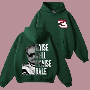 May include: A dark green hoodie featuring a graphic of a man in sunglasses with the words "RAISE HELL PRAISE DALE" in white. The back of the hoodie has a red and white number 3 logo. The hoodie has a hood and a front pocket.