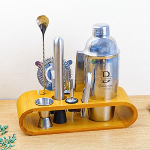 Pode incluir: Um conjunto de coqueteleira de aço inoxidável com uma base de madeira de bambu. O conjunto inclui uma coqueteleira, um medidor, uma colher de bar, um pilão, um coador e outras ferramentas de bar. A coqueteleira é gravada com o nome "Brooklyn" e a palavra "Groomsman".