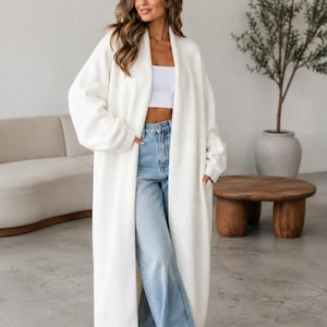 May include: A long, open-front cream-colored cardigan with long sleeves and side pockets, worn over a white cropped top and light blue denim jeans. The cardigan features a smooth, solid-colored knit texture and falls to the lower calf area.
