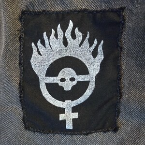 May include: Black patch with a silver graphic of a skull within a circle, flames, and a cross symbol. The patch is sewn onto a gray denim fabric, with visible stitching around the edges. The design has a distressed, punk rock aesthetic.