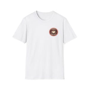 Op de afbeelding: Een wit T-shirt met een ronde hals en korte mouwen. Een cirkelvormige afbeelding op de borst toont een kat in een koffiekopje met de woorden "MAMA NEEDS COFFEE" in een bruin en rood ontwerp.