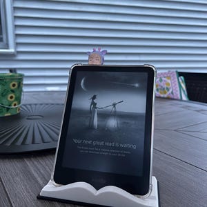 May include: A black Kindle e-reader in a clear protective case, resting on a white book-shaped stand. The screen displays a black and white image of a person using a telescope under a crescent moon, with the text Your next great read is waiting.