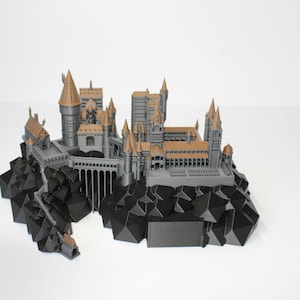 May include: A detailed model of a castle with multiple towers and structures, in shades of gray and gold. The castle sits atop a black, geometric rock formation. The model is likely a replica of a famous castle.