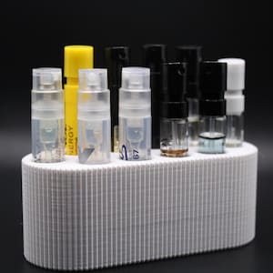 May include: A white, ribbed, oval-shaped perfume sample holder with a variety of perfume vials. The vials are clear, black, yellow, and white, with some containing liquid. The background is black.