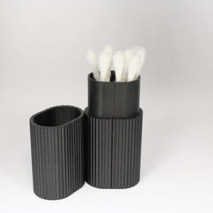 May include: A black, ribbed, cylindrical container with a matching lid, holding several white cotton swabs. The container is open, revealing the swabs. The lid is placed next to the container.
