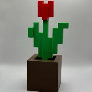 Könnte beinhalten: Eine 3D-gedruckte dekorative Tulpe in einem braunen quadratischen Topf. Die Tulpe hat eine rote Blüte und einen grünen Stiel und Blätter. Das Design ist pixelig und verleiht ihm eine Retro-Videospiel-Ästhetik.