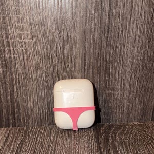 Könnte beinhalten: Ein weißes Ladecase für kabellose Kopfhörer mit einer glatten, abgerundeten rechteckigen Form. Ein dünnes, leuchtend rosa Silikonband ist horizontal um die Mitte des Gehäuses gewickelt und bildet einen deutlichen Kontrast zur weißen Oberfläche.